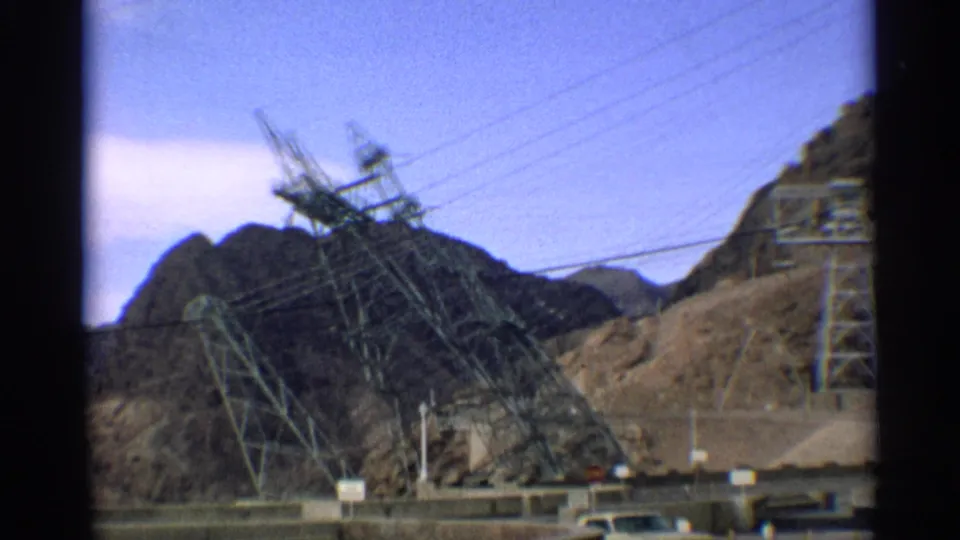 1959 Arizona 8mm frame with transmission towers and high-voltage lines set against rugged desert mountains near a dam access road.