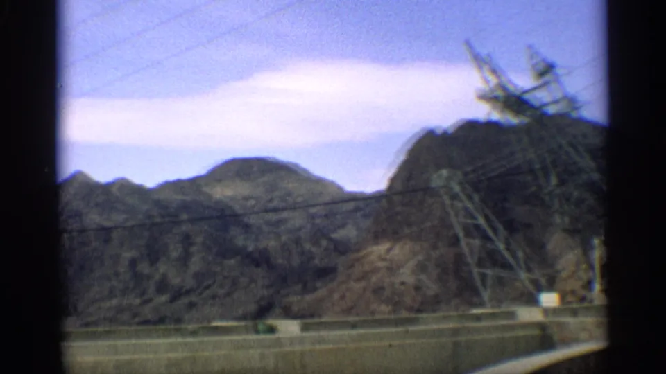 1959 Arizona 8mm frame showing rugged desert mountains, utility lines, a transmission tower, and part of a concrete structure from a moving roadside view.