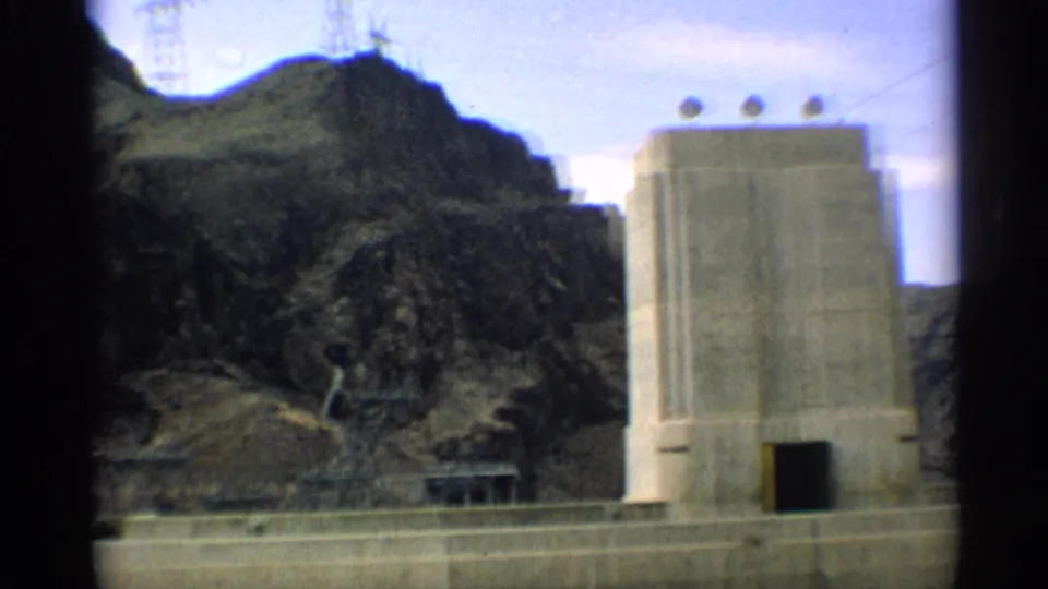 1959 Arizona 8mm frame showing the concrete structure from a passing vehicle, with arid slopes, utility lines, and a small roadside building or shelter.