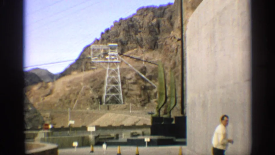 1959 Arizona frame showing a large concrete structure beside utility equipment, with an adult man standing in the foreground.