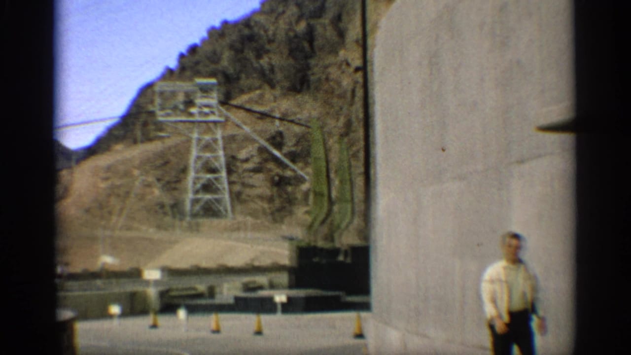 Man standing near a curved concrete dam structure with mountains and power equipment behind him.