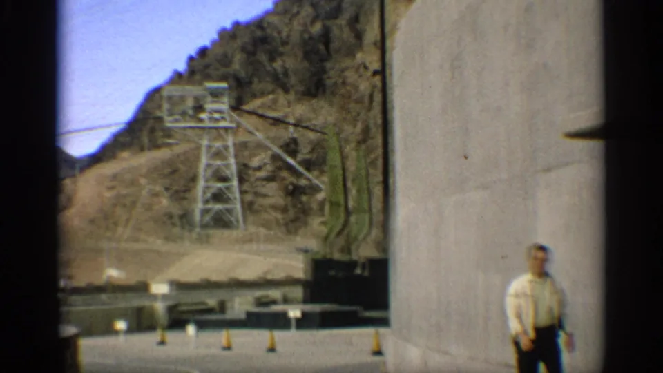 In 1959 Arizona footage, an adult man stands near a curved concrete dam wall with rugged hills, road cones, and power equipment in the background.