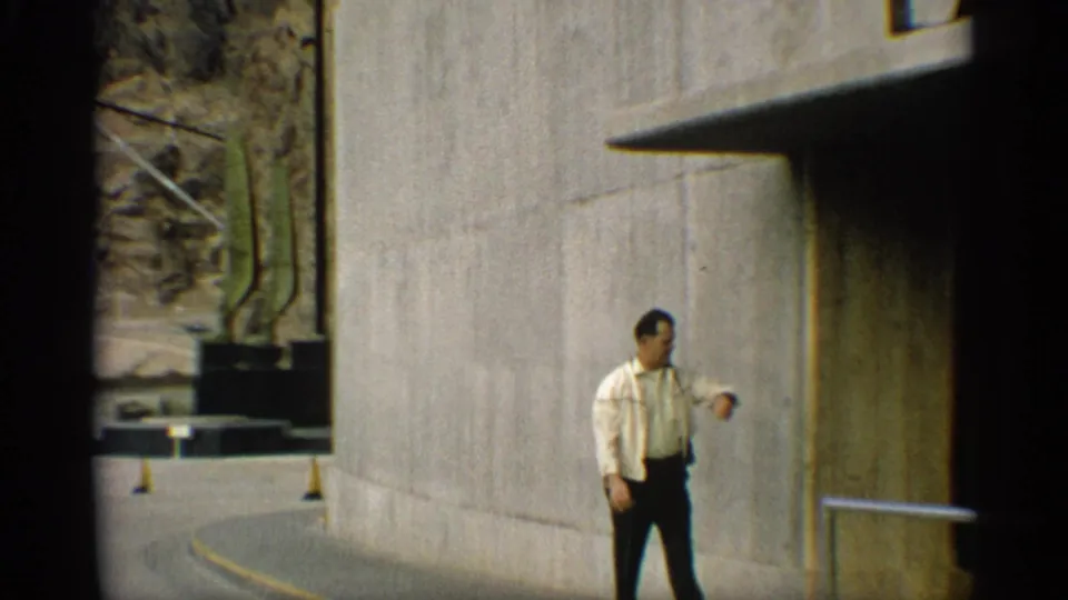 An adult man in a light shirt and dark pants walks beside a tall concrete dam wall along a curved roadway in the Arizona setting.