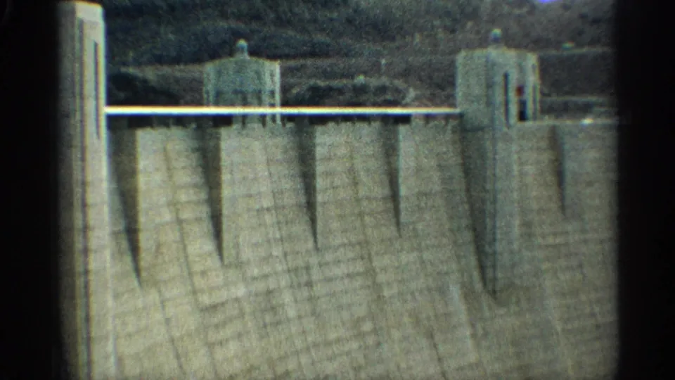 The footage cuts to a large concrete dam face with vertical tower structures and rocky hills behind it, seen in soft 8mm color.