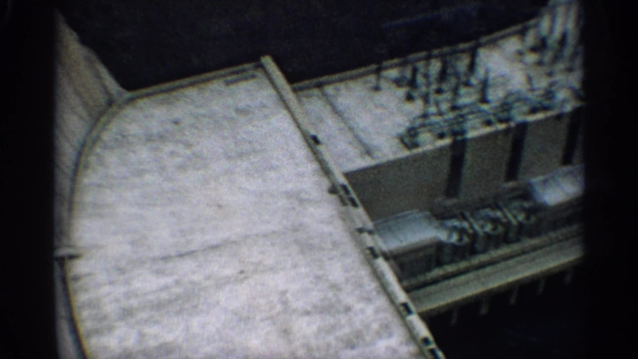 1959 Arizona 8mm frame showing an overhead view of concrete dam construction with equipment lined along a service deck.