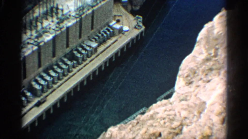 1959 Arizona frame showing dam construction equipment along a concrete edge with dark water and pale canyon rock nearby.
