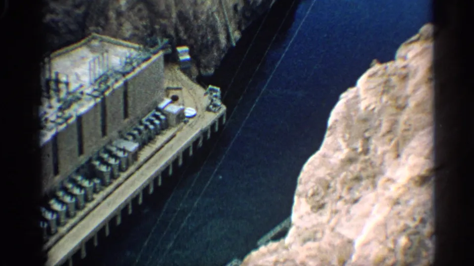Aerial 8mm frame of Arizona dam construction, with concrete structures, water, and rugged canyon rock in view.