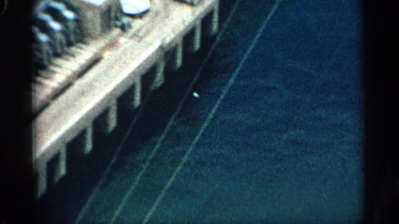 1959 Arizona 8mm frame with an oblique aerial view across a concrete dam edge, reservoir water, and pale canyon rock.