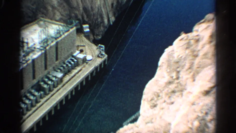 1959 Arizona 8mm frame looking down on dam equipment, crossing utility lines, blue canyon water, and a bright rock face.