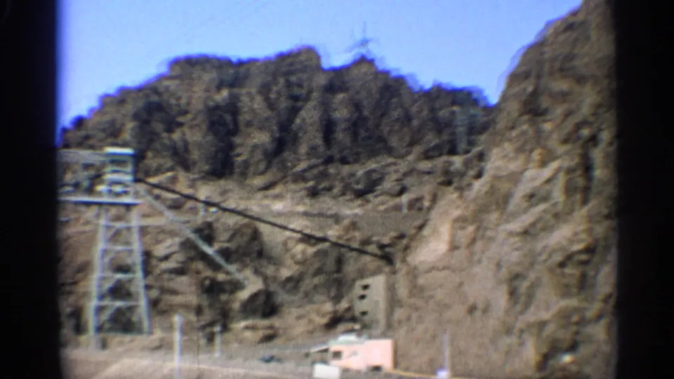 1959 Arizona 8mm frame showing a rocky canyon slope, utility lines, a tower-like structure, and a small pale building near the dam area.