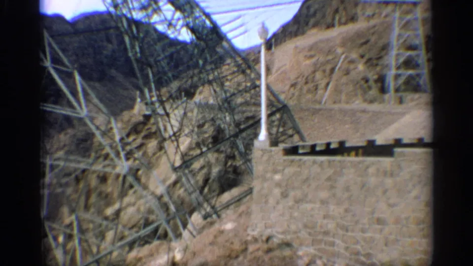 1959 Arizona 8mm frame with angled metal transmission structures, overhead lines, a stone retaining wall, and arid hillside terrain.
