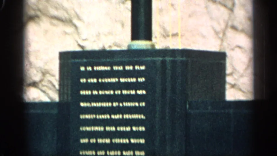 1959 Arizona 8mm frame showing a closer view of a dark stone monument plaque with engraved text and a rough wall behind it.