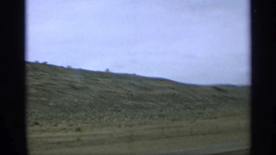 1959 Arizona travel frame showing a low dry hillside and pale sky from a moving vehicle, with soft 8mm motion blur.