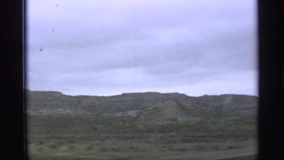 1959 Arizona home-movie frame showing rolling green hills under a cloudy sky from a moving road-trip viewpoint.