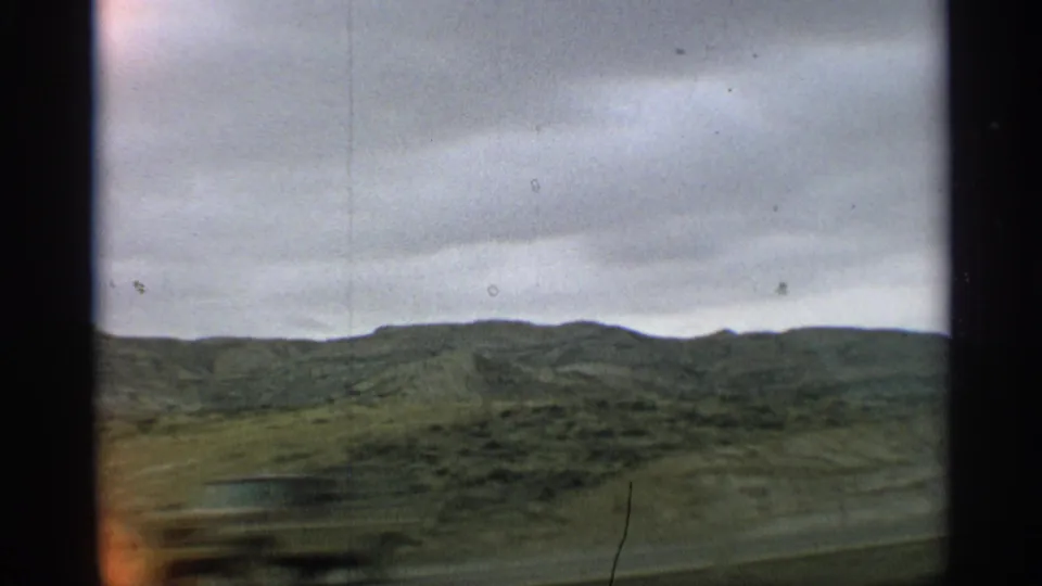1959 Arizona travel frame showing green hills under clouds with blurred roadside foreground from a moving car.