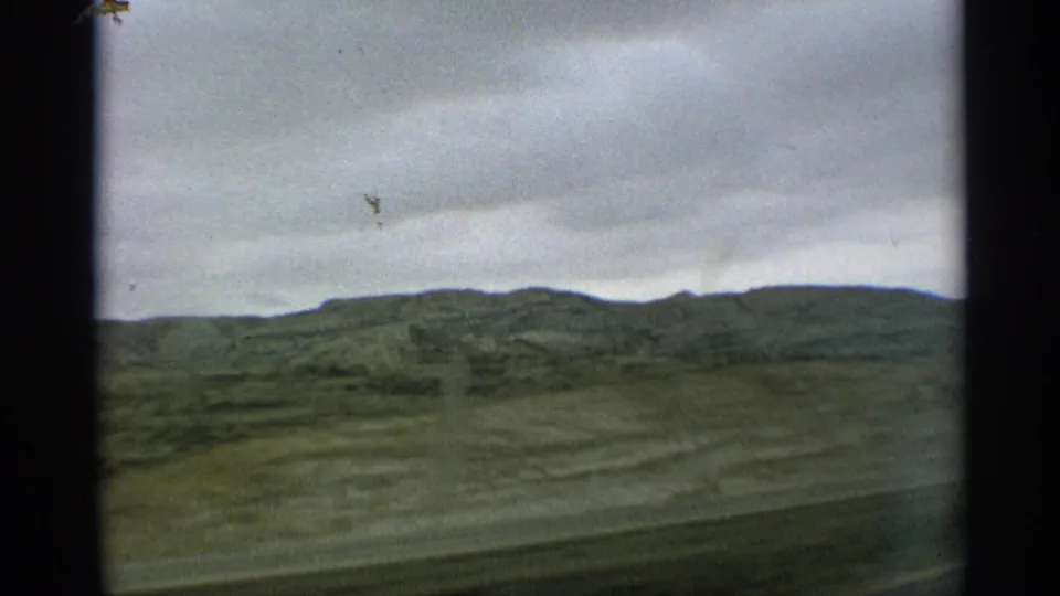 1959 Arizona 8mm travel frame showing grassy hills beneath a gray cloud layer, viewed from a moving vehicle.