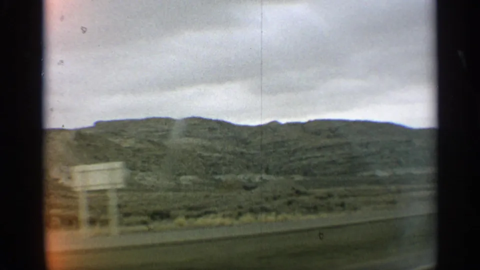 1959 Arizona roadside landscape with green hills, overcast sky, and a brief pale sign or structure shape near the foreground.