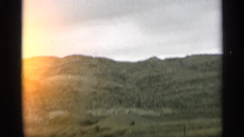 1959 Arizona travel view of grassy hills under a cloudy sky, softened by vehicle motion and warm edge flare on the 8mm frame.