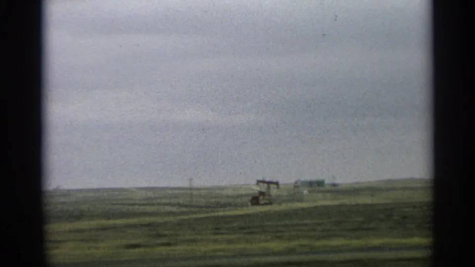 1959 Arizona 8mm frame showing a distant oil pump jack across a green field under a cloudy sky.