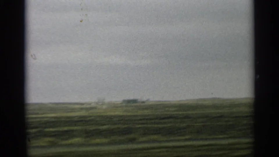 1959 Arizona 8mm frame of a blurred green field and distant low structure seen from a moving vehicle.