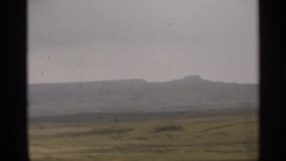 Soft 8mm frame of Arizona desert fields with low mountains in haze, filmed from a moving vehicle in 1959.