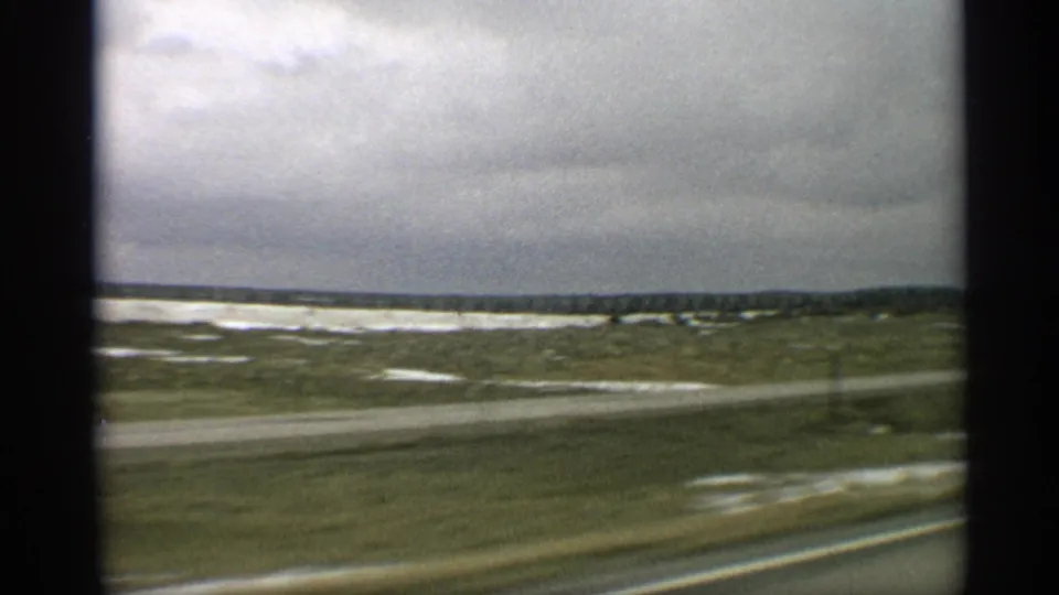 Late frame from the 1959 Arizona car view, showing roadside blur, open desert, pale patches, and distant low landforms under clouds.