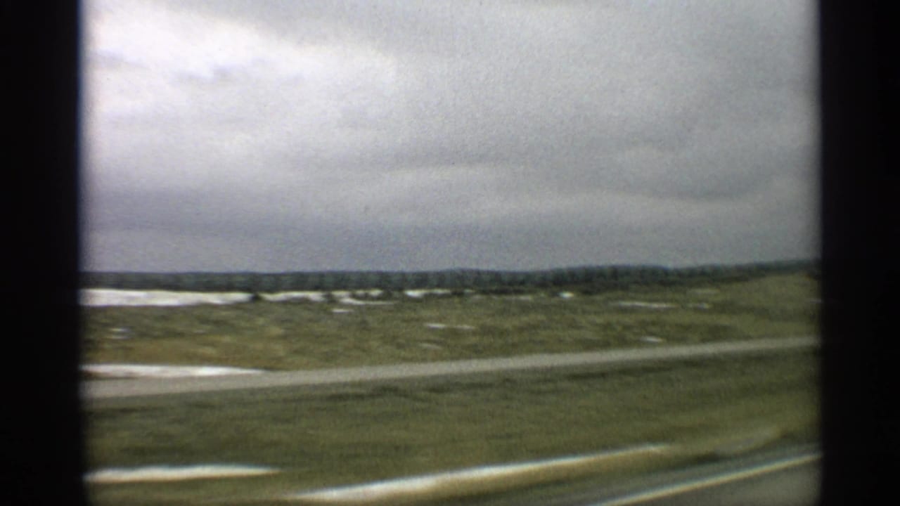 1959 Arizona 8mm frame showing a moving view past open rural fields, pale ground patches, and a cloudy sky.