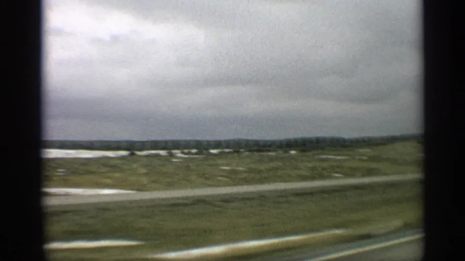 1959 Arizona 8mm frame showing a moving view past open rural fields, pale ground patches, and a cloudy sky.