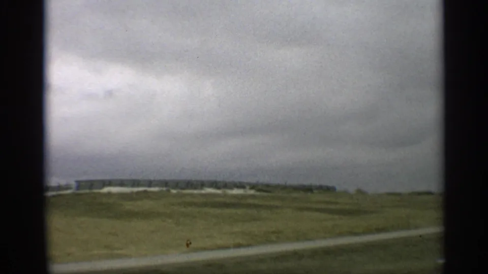 1959 Arizona home-movie frame with a low building or structure beyond open land and a pale road edge under cloudy sky.