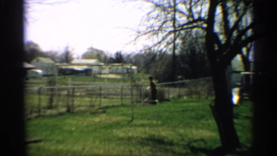 1959 Arizona home-movie frame showing a small childlike figure in a grassy backyard near a chain-link fence, with houses and bare trees behind.