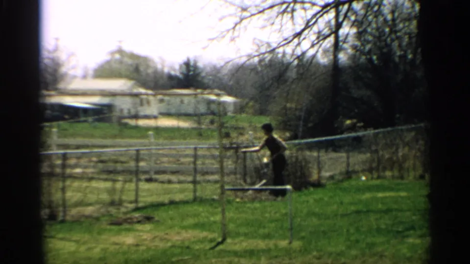 In 1959 Arizona home-movie footage, a woman works near a backyard fence with grass, bare branches, and a distant white house visible.