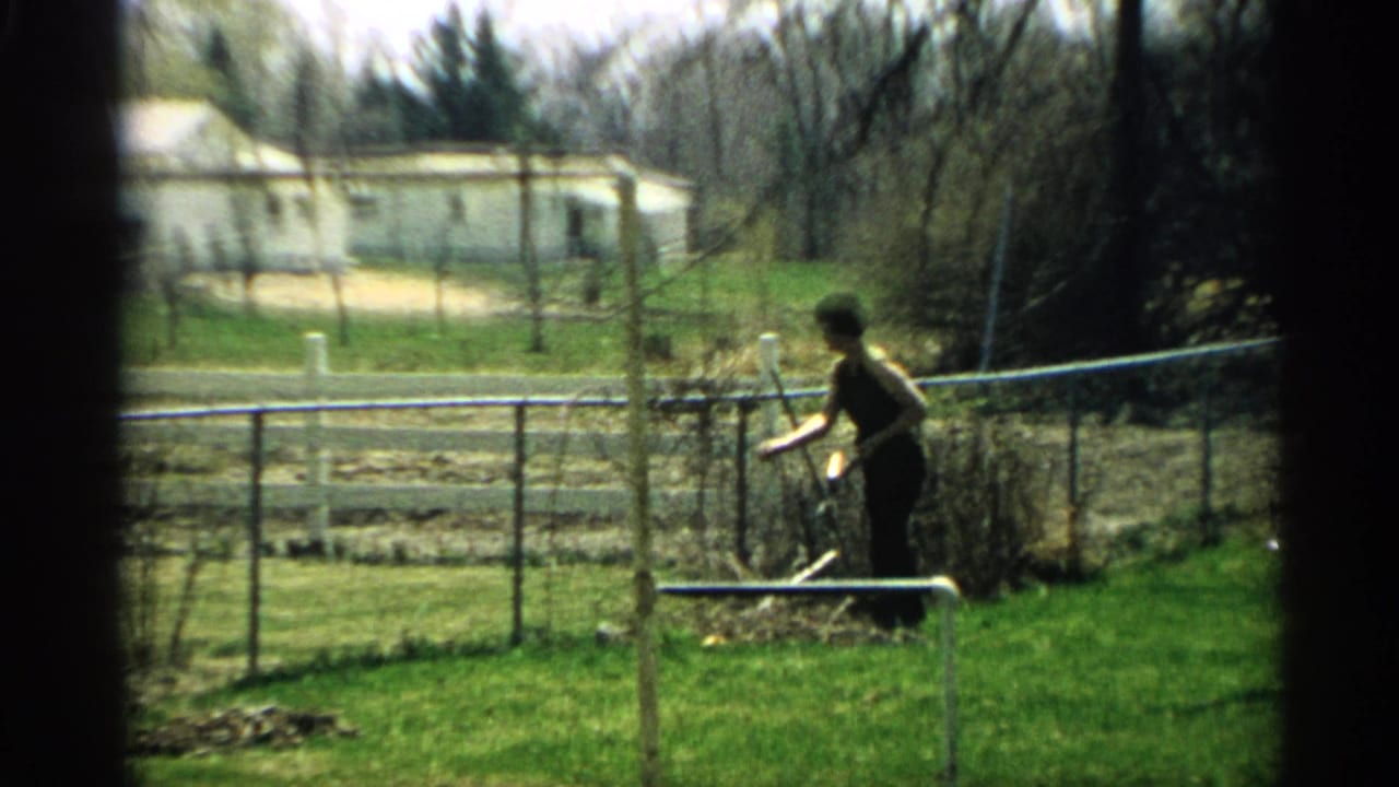Woman holding a small tool beside a fence, with garden beds and a house in the background.