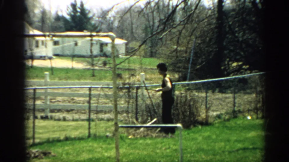A woman in dark clothing stands beside a chain-link fence, with lawn, small garden rows, and a light-colored house behind her.