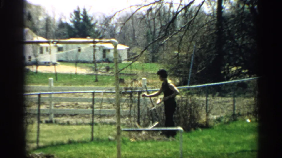 At the end of the 1959 Arizona clip, the woman remains near the fence in the backyard, with trees and a house beyond the lawn.