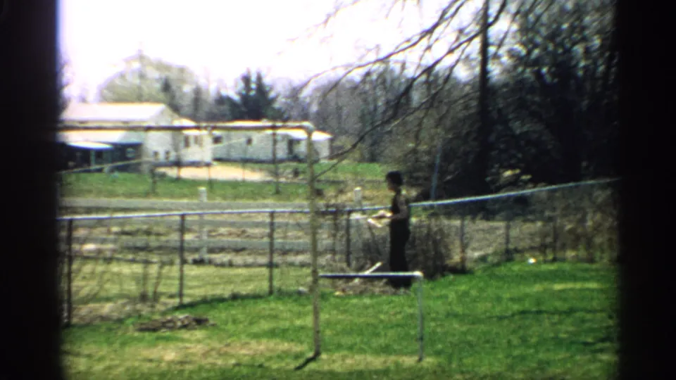 The person continues yard work near the chain-link fence, with garden rows, trees, and a mobile-home-like structure in the background.