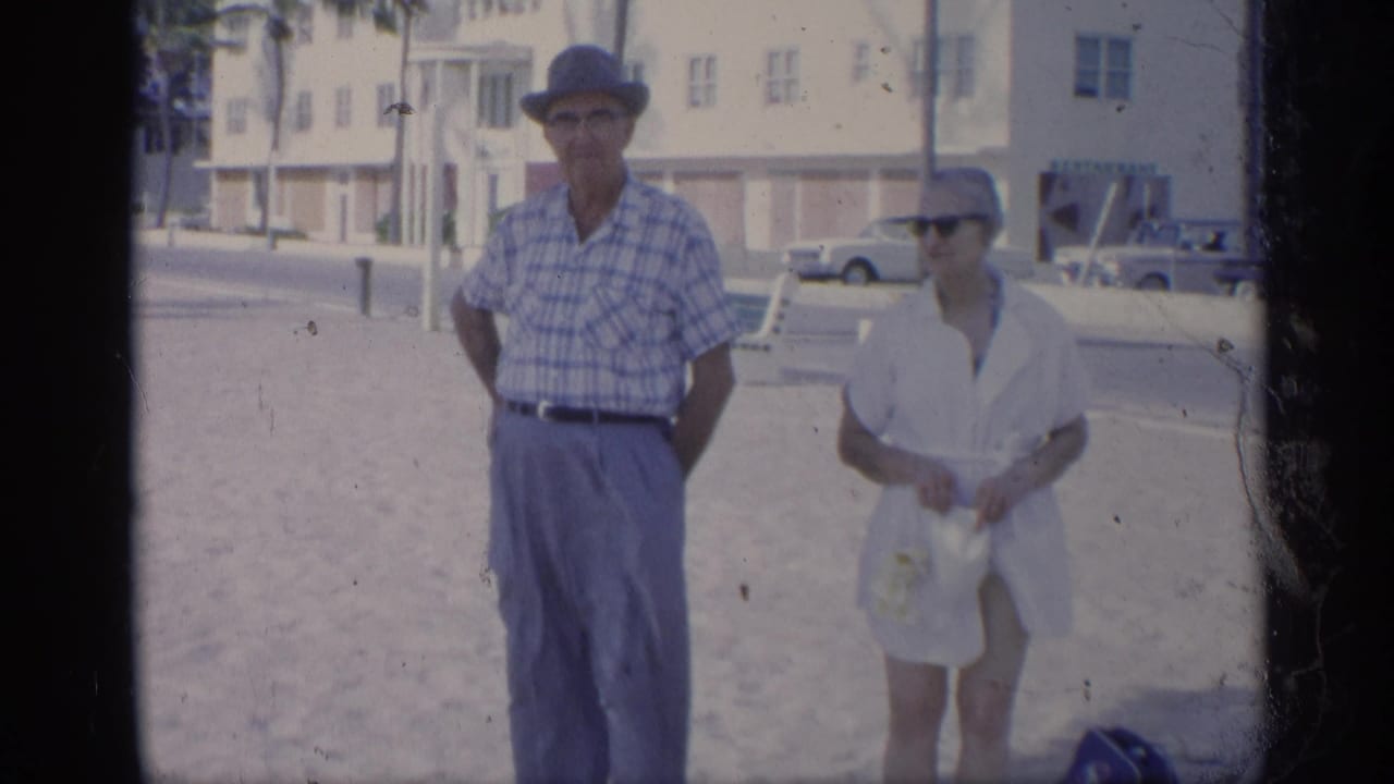 Man in plaid shirt and woman in white cover-up stand on a sunny beach.