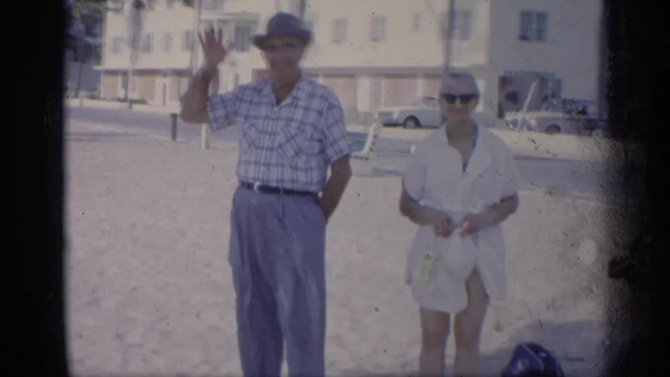 The man waves toward the home-movie camera while the woman stands beside him on a sunny California beach.