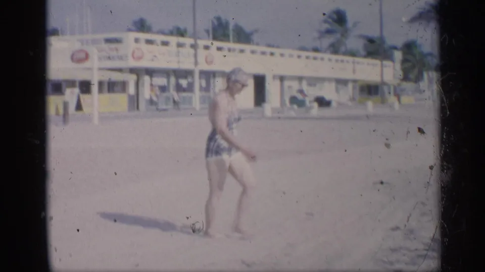 A woman in a patterned swimsuit walks on the sandy beach, with palm trees and a low seaside building behind her.