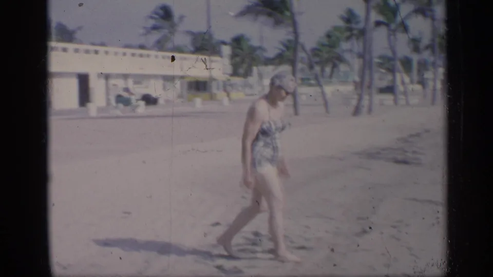 The woman continues walking across the sunlit California beach, framed by palms, sand, and a seaside building.
