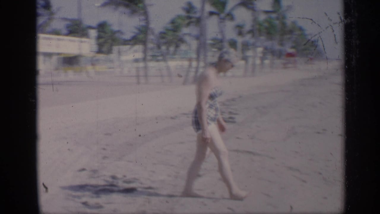 Woman in a plaid swimsuit walking barefoot on a sandy beach with palm trees behind her.