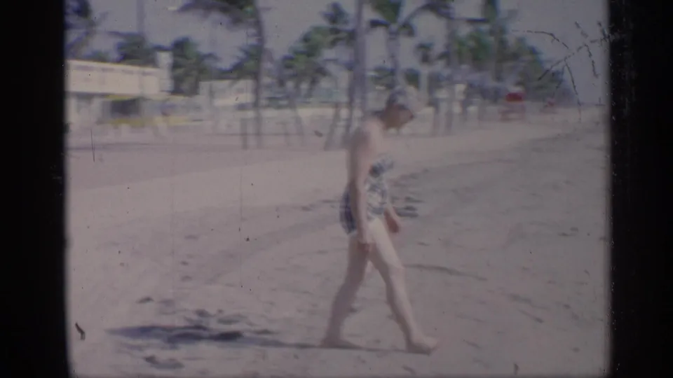 1959 California home-movie frame of a woman in a plaid one-piece swimsuit and swim cap walking along a sandy beach.