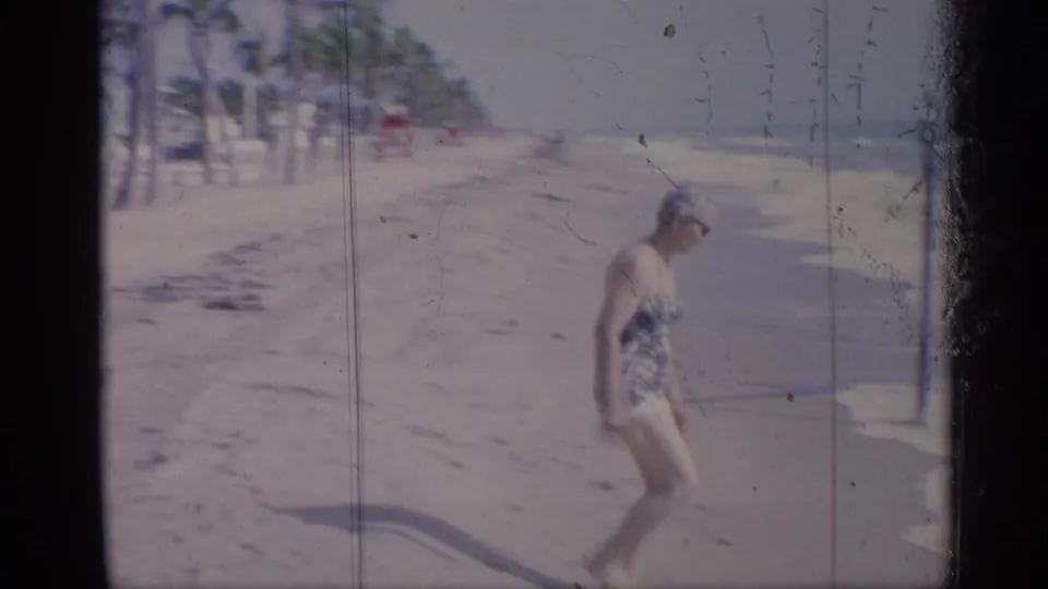 Woman in plaid swimwear walks along a sunlit California beach in 1959, seen in soft 8mm home-movie footage.