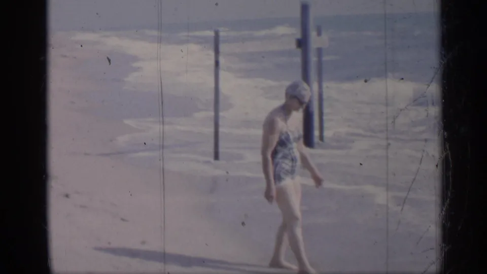 1959 California beach frame showing a woman in a plaid one-piece swimsuit walking near posts and foamy surf.