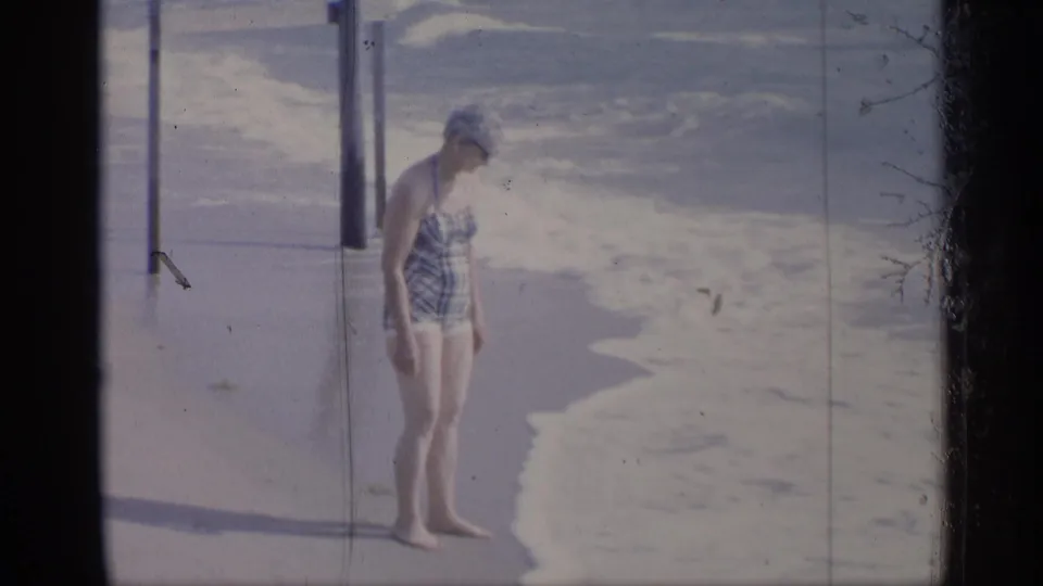 Woman in plaid swimwear stands by the foamy shoreline in a 1959 California 8mm beach home movie frame.