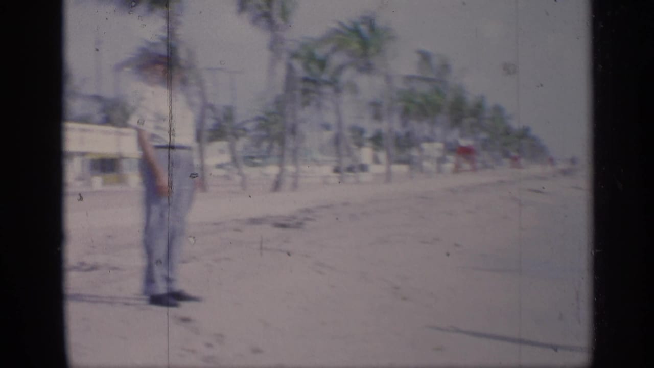 A dressed man stands on a sandy beach with palm trees and a low building behind him.