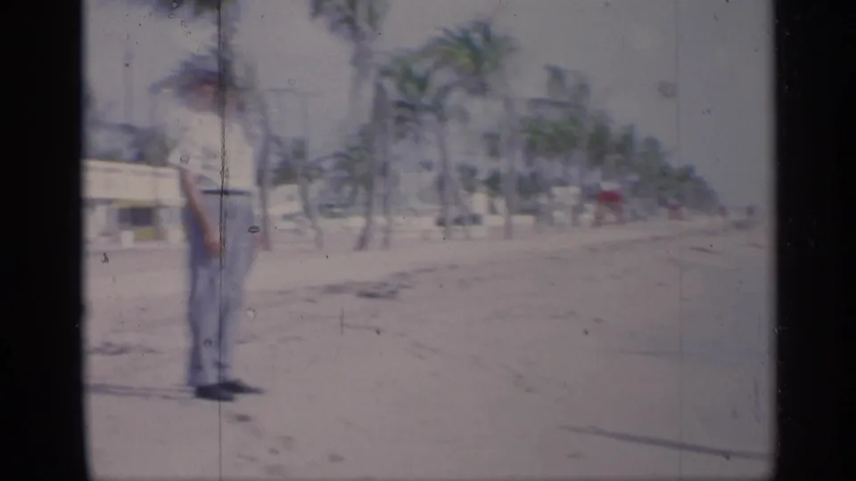 A man in light clothing and a hat stands on a California beach in 1959, with palm trees and a beachside building in the background.