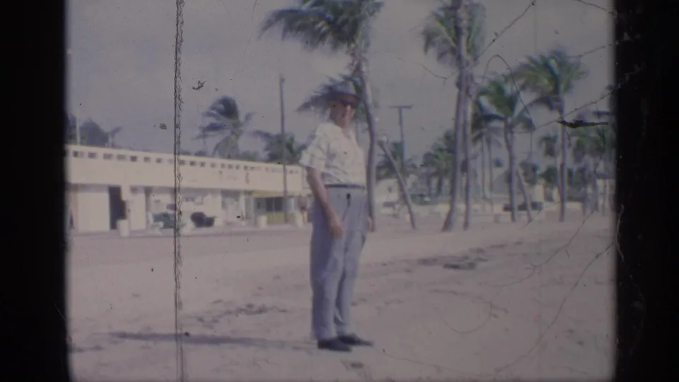 A well dressed man in a hat stands on a 1959 California beach, with windblown palms and a low building behind him.