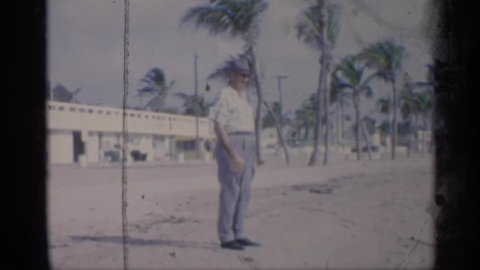 A man in light shirt, trousers, sunglasses, and hat stands on a California beach in 1959 near windblown palm trees.