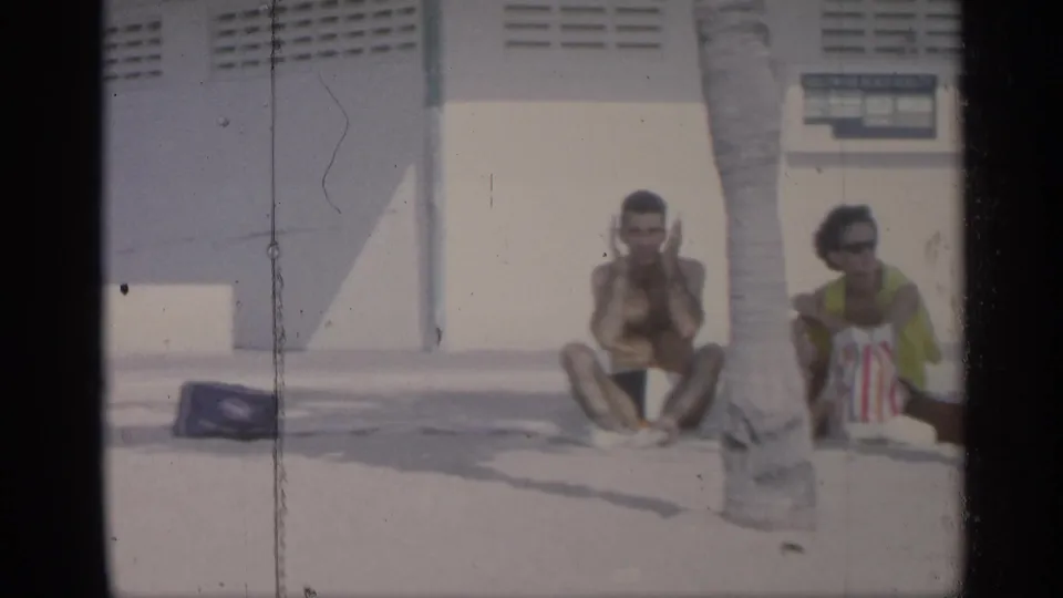 Two adults relax near a palm tree and light-colored beachside building in the 1959 California home-movie footage.