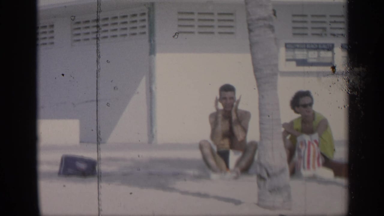 Two beachgoers sit near a palm tree beside a vented beach building.